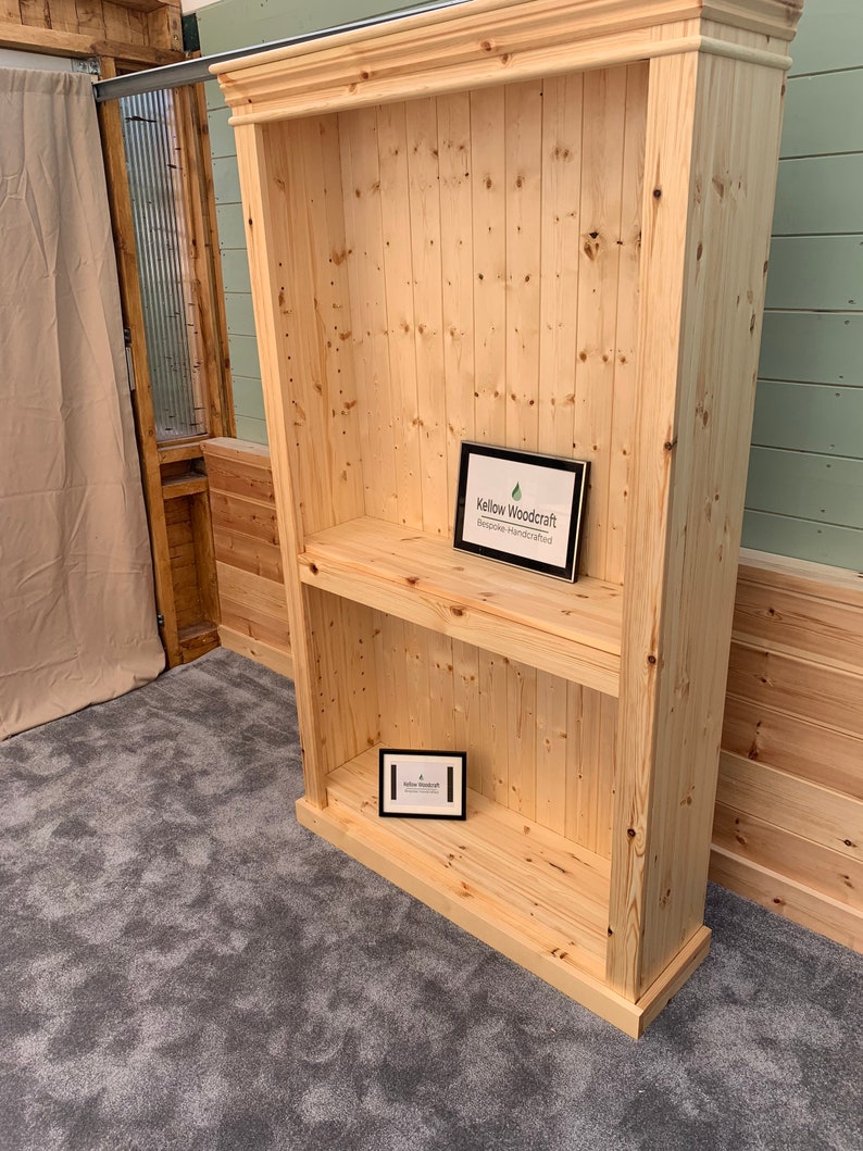 May include: A tall, wooden bookcase with two shelves. The bookcase is made of natural wood and has a simple, rustic design. The bookcase is empty, but there is a small sign on the bottom shelf that says "Kellow Woodcraft - Bespoke Handcrafted".