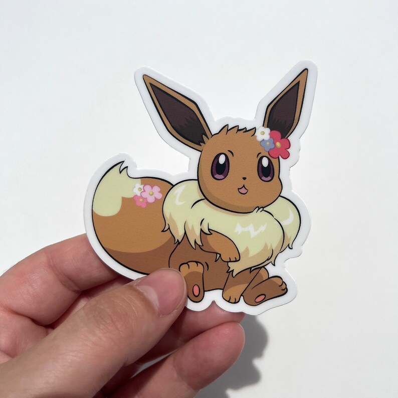 May include: A brown and cream colored Eevee Pokemon sticker with a flower crown. The Eevee is sitting with its tail curled around its body. The sticker is on a white background.