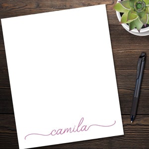 May include: A white sheet of paper with the word "camilla" written in cursive in pink on the bottom. The paper is on a wooden surface with a potted succulent and a black pen.