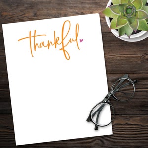 May include: A white notepad with the word "thankful" written in orange cursive with a small pink heart next to it. A pair of black eyeglasses and a potted succulent plant are on a wooden surface.