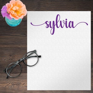 May include: A white sheet of paper with the name "sylvia" written in purple cursive script. A pair of black-framed glasses and a vase of flowers are on a wooden surface.