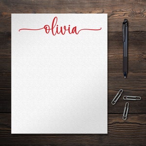 May include: A white sheet of paper with the name "Olivia" written in red cursive script on the top. The paper is on a dark brown wooden surface with a black pen and three silver paper clips.