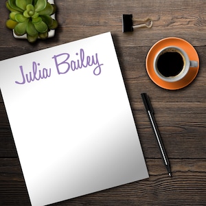 May include: A white sheet of paper with the name "Julia Bailey" written in purple cursive on the top. The paper is on a wooden table with a cup of coffee, a pen, and a potted succulent.