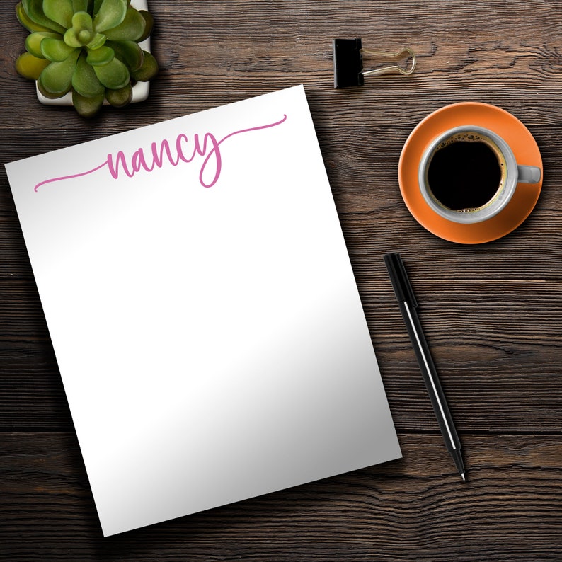 May include: A white notepad with the name "nancy" written in pink cursive on the top. The notepad is on a wooden surface with a cup of coffee, a pen, and a paperclip.