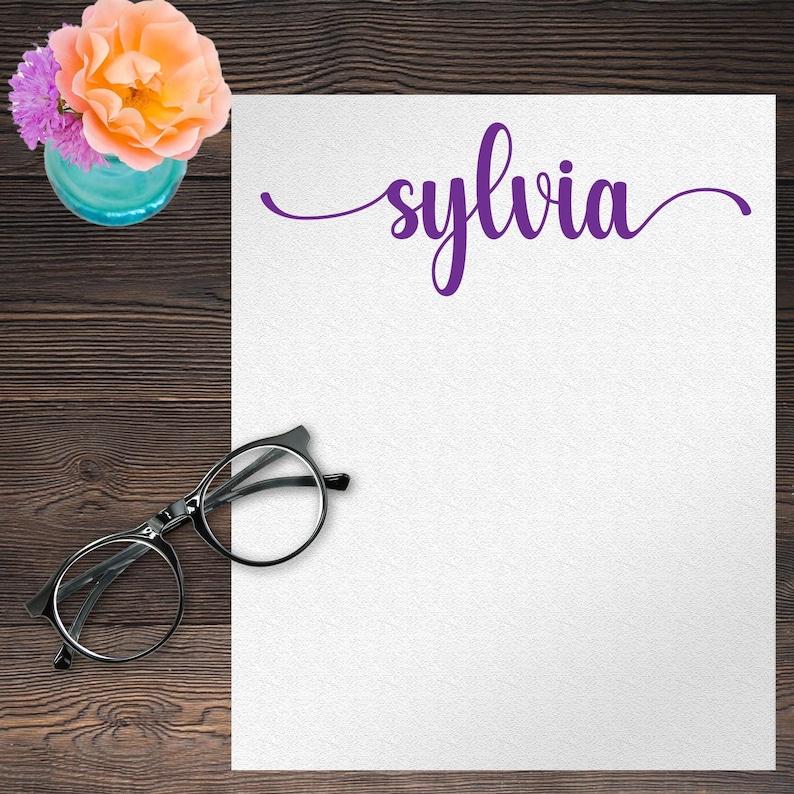 Pode incluir: Uma folha de papel branca com o nome "sylvia" escrito em letra cursiva roxa. Um par de &oacute;culos com arma&ccedil;&atilde;o preta e um vaso de flores est&atilde;o sobre uma superf&iacute;cie de madeira.