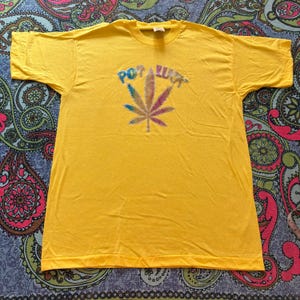 May include: A yellow t-shirt with a colorful cannabis leaf design and the words "Pot Luck" in a rainbow font. The shirt is laid flat, showcasing its design and color.