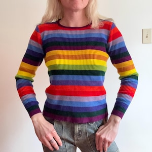 May include: A long-sleeved, ribbed knit sweater with horizontal stripes in a rainbow of colors. The sweater has a crew neck and a close fit. The colors include red, orange, yellow, green, blue, and purple. The sleeves are long and fitted.