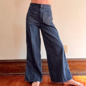 May include: Dark blue denim wide-leg pants with decorative stitching on the back pockets. The pants have a high waist and a button closure. The fabric appears to be a medium weight denim, suitable for various seasons.
