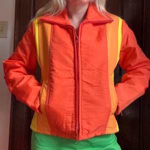 May include: A vibrant orange jacket with a red zipper and yellow side stripes. The jacket has a collared neckline and two front pockets. The sleeves are long and the jacket is worn over green pants. A retro style jacket.
