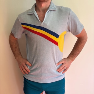 May include: Light blue polo shirt with a V-neck collar and short sleeves. The shirt features a diagonal stripe design in red, yellow, and blue, with a yellow curved shape on the right side. The shirt is paired with teal corduroy shorts.