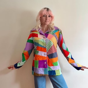 May include: A patchwork jacket with a variety of colorful fabric squares in shades of red, orange, green, purple, and blue. The jacket has a button-up front and a purple collar. The sleeves are long and the jacket is worn with blue jeans.