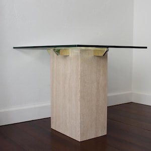 May include: A square glass-top side table with a light beige, rectangular base. The base is made of a stone-like material. The table is set against a white wall and dark wood floor.