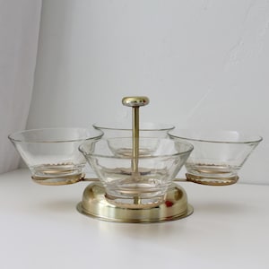 May include: A revolving serving set featuring four clear glass bowls arranged around a central brass-toned metal post. The bowls are supported by a circular brass-toned base with a matching central handle, creating a tiered structure for food.