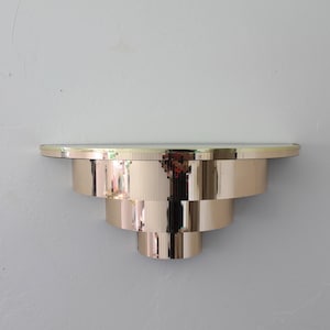 May include: A wall-mounted shelf featuring a tiered, stepped design with a polished, reflective rose gold metallic finish. The structure consists of three concentric circular layers that decrease in size toward the bottom, topped with a flat surface.