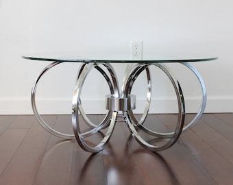 May include: A round glass top coffee table with a chrome base featuring four interlocking rings. The table is on a dark wood floor.