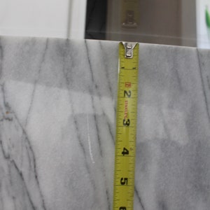 May include: A close-up of a white marble countertop with a yellow measuring tape placed vertically against it. The tape measures 7 inches.