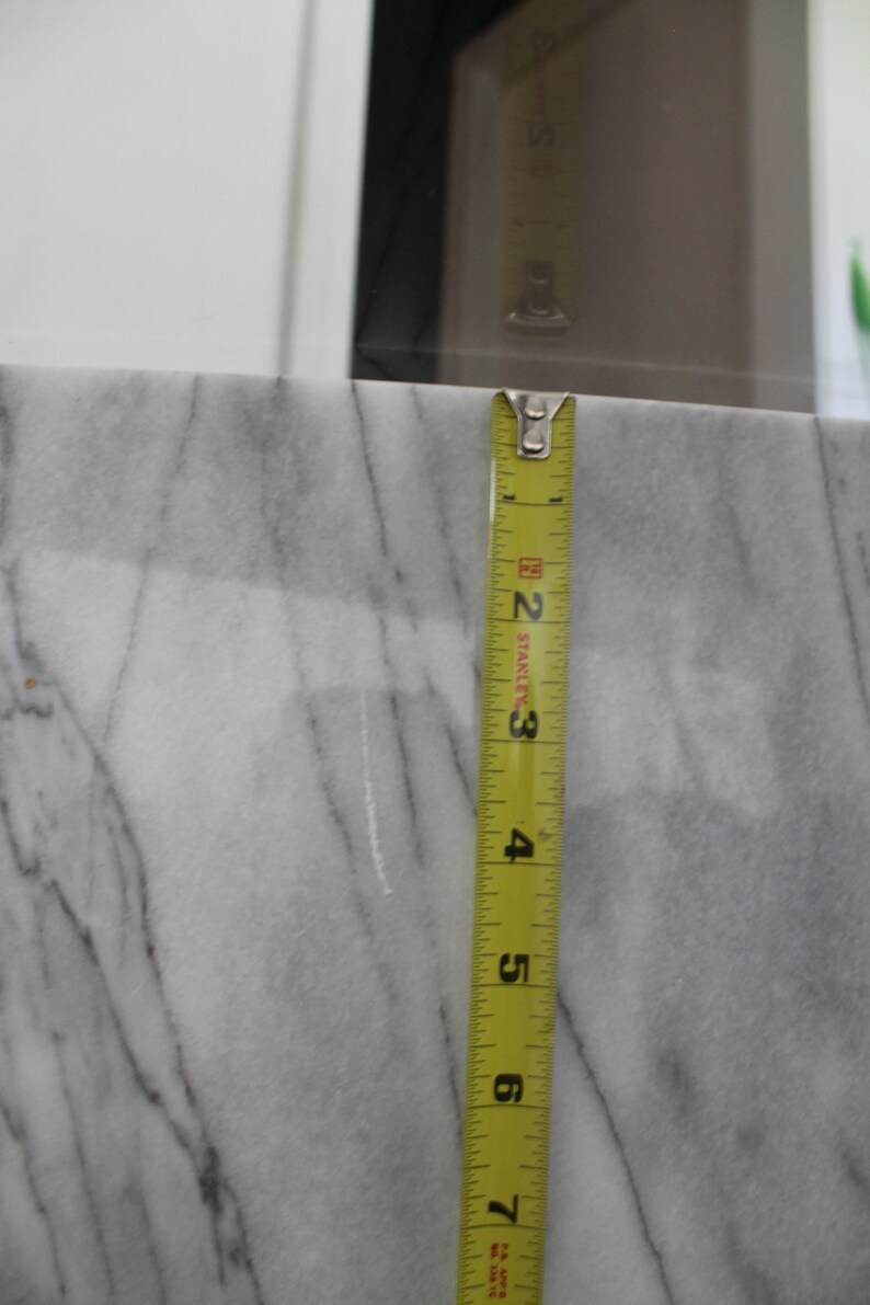 May include: A close-up of a white marble countertop with a yellow measuring tape placed vertically against it. The tape measures 7 inches.