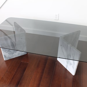 May include: A rectangular glass-top coffee table with a smoked glass top and a white marble base. The base is made of two pieces of marble that are joined together in a unique way.