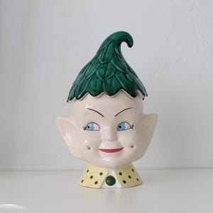 May include: A ceramic elf-shaped cookie jar with a green leaf-like hat. The face is painted with blue eyes, a red mouth, and rosy cheeks. The collar is yellow with green polka dots and a green button.