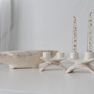 May include: A cream-colored ceramic bowl and two matching candle holders with gold leaf accents. The candle holders each hold two candles with gold leaf embedded in the clear wax. The bowl has a speckled design.