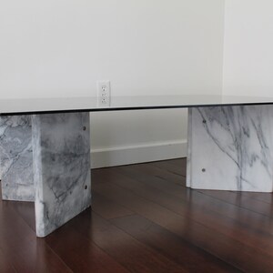 May include: A rectangular glass-top coffee table with a white marble base. The table has four legs, each made of two pieces of marble that are joined together in a V-shape.