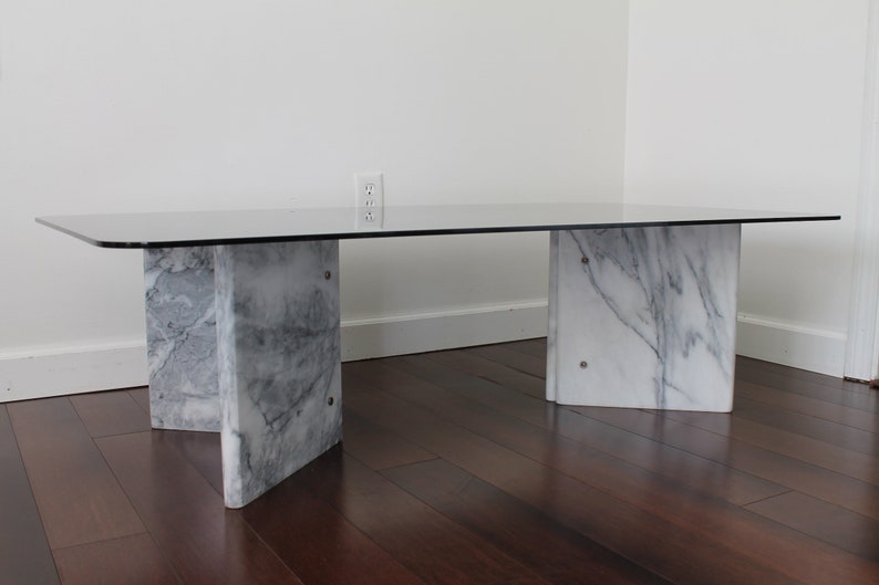 May include: A rectangular glass-top coffee table with a white marble base. The table has four legs, each made of two pieces of marble that are joined together in a V-shape.