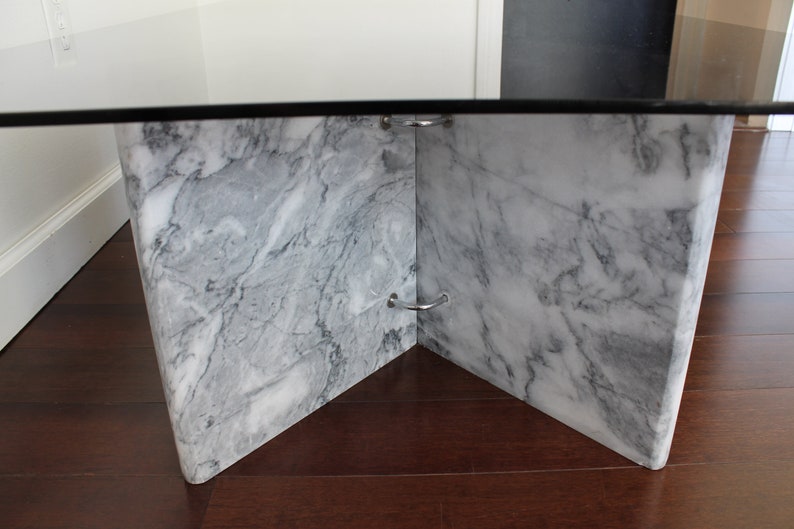 May include: A glass-top coffee table with a white marble base. The base is made of four pieces of marble that meet in the center, creating a unique and modern design.