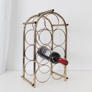 May include: A gold-colored metal wine rack with a bamboo-style design. The rack holds multiple bottles and features circular openings. A single bottle of red wine is placed in the rack, showcasing its storage capacity and decorative appeal.