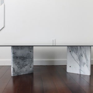 May include: A modern coffee table with a clear glass top and two white marble bases. The table is in a room with hardwood floors and white walls.