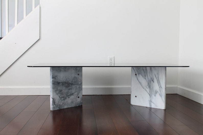 May include: A modern coffee table with a clear glass top and two white marble bases. The table is in a room with hardwood floors and white walls.