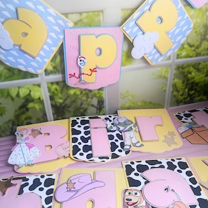 Peut inclure: Une guirlande d&eacute;corative avec des lettres roses et jaunes, un motif de vache et des d&eacute;coupes de personnages. Les personnages incluent une princesse, un cochon, Buzz l'&Eacute;clair, un chapeau de cow-boy et un chien. La guirlande est d&eacute;cor&eacute;e de nuages et d'&eacute;toiles.
