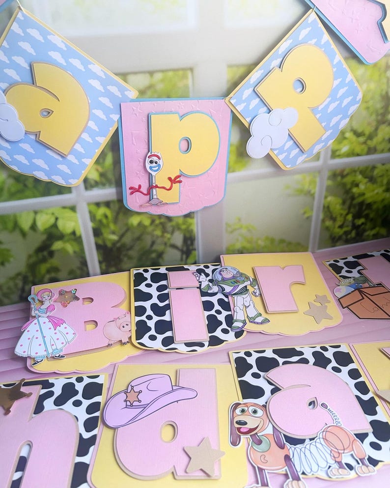 Peut inclure: Une guirlande d&eacute;corative avec des lettres roses et jaunes, un motif de vache et des d&eacute;coupes de personnages. Les personnages incluent une princesse, un cochon, Buzz l'&Eacute;clair, un chapeau de cow-boy et un chien. La guirlande est d&eacute;cor&eacute;e de nuages et d'&eacute;toiles.