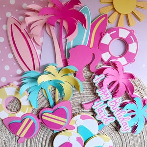 Op de afbeelding: Een verzameling kleurrijke papieren uitsparingen met een zomerthema. Bevat palmbomen, surfplanken, zonnebrillen, strandballen, een zon en een reddingsboei. De woorden "Happy Birthday Hazel" zijn ook aanwezig. Het kleurenpalet omvat roze, geel en blauw.