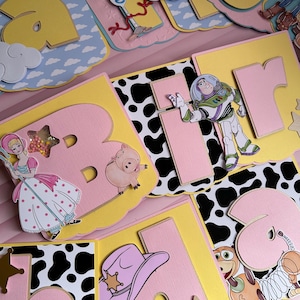 Peut inclure: Une banni&egrave;re color&eacute;e "Joyeux Anniversaire" avec des personnages de Toy Story. La banni&egrave;re comprend des images de Buzz l'&Eacute;clair, Woody, Bo Peep et d'autres personnages, avec un code couleur rose, jaune et bleu.
