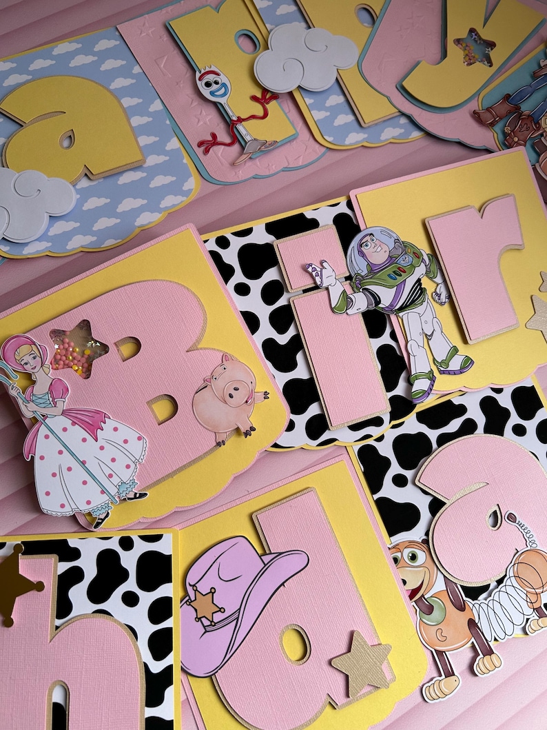 Peut inclure: Une banni&egrave;re color&eacute;e "Joyeux Anniversaire" avec des personnages de Toy Story. La banni&egrave;re comprend des images de Buzz l'&Eacute;clair, Woody, Bo Peep et d'autres personnages, avec un code couleur rose, jaune et bleu.