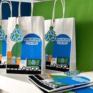 Może przedstawiać: Białe papierowe torby prezentowe z zielonymi etykietami z symbolem recyklingu i wzorem niebieskiego i zielonego śmieciarki. Torby mają napis "Dzięki, że przyszedłeś na imprezę!"