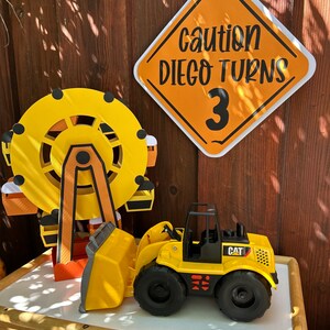 Puede incluir: Un vehículo de construcción de juguete amarillo con un logotipo "CAT" negro, una noria de juguete amarilla y una señal de construcción amarilla y negra que dice "Precaución Diego cumple 3 años".