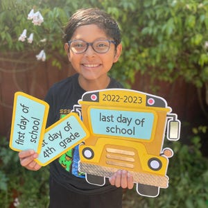 May include: A young person holding signs that say "first day of school" and "last day of 4th grade". The signs are yellow with blue text. The person is also holding a yellow school bus cutout with the text "2022-2023 last day of school".