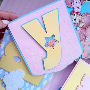 Peut inclure: Une page de scrapbooking faite &agrave; la main avec une grande lettre "Y" jaune et une fen&ecirc;tre en forme d'&eacute;toile remplie de confettis color&eacute;s. La page a un fond rose avec des &eacute;toiles en relief et des bordures bleues. Une image de Woody de Toy Story est visible.