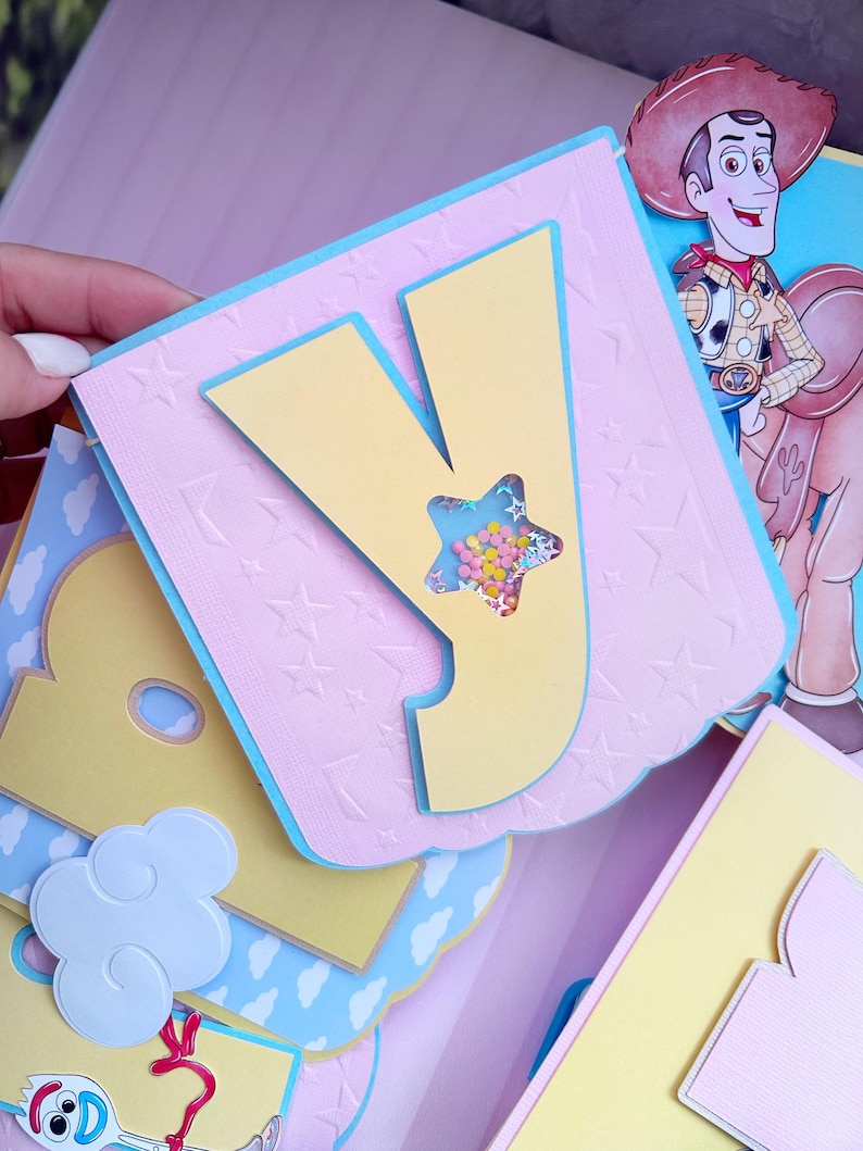 Peut inclure: Une page de scrapbooking faite &agrave; la main avec une grande lettre "Y" jaune et une fen&ecirc;tre en forme d'&eacute;toile remplie de confettis color&eacute;s. La page a un fond rose avec des &eacute;toiles en relief et des bordures bleues. Une image de Woody de Toy Story est visible.
