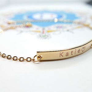 Op de afbeelding: Een goudkleurige armband met een fijne ketting en een gebogen staaf. De staaf is gegraveerd met de naam "Katie" en een hartsymbool. De armband is ontworpen voor dagelijks gebruik.