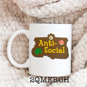 May include: White ceramic mug with a brown sign that says "Anti-Social" with a green leaf and a peach on it. The sign is in the style of a "Welcome to" sign from the video game Animal Crossing. The mug is on a white fluffy surface.