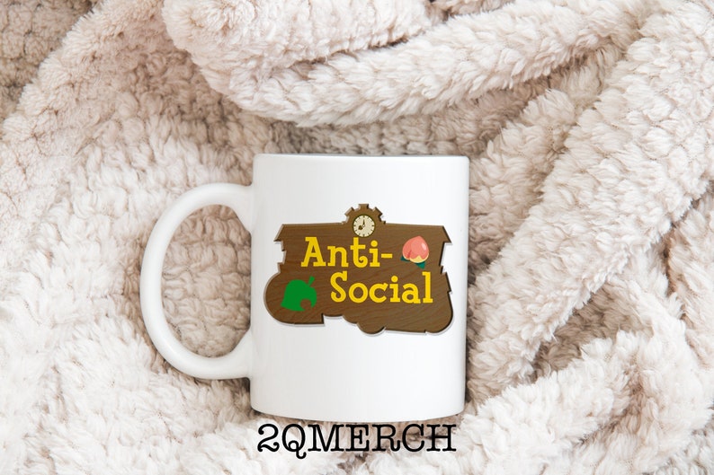 May include: White ceramic mug with a brown sign that says "Anti-Social" with a green leaf and a peach on it. The sign is in the style of a "Welcome to" sign from the video game Animal Crossing. The mug is on a white fluffy surface.