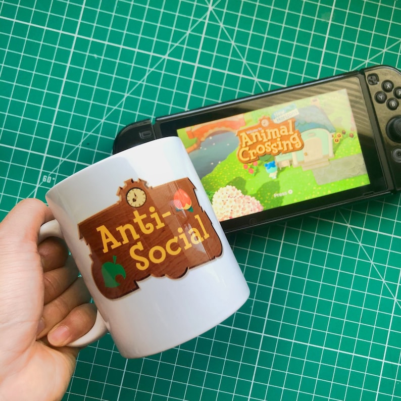 May include: White ceramic mug with a brown sign that says "Anti-Social" with a green leaf and a peach on it. The mug is next to a Nintendo Switch with the Animal Crossing game on the screen.