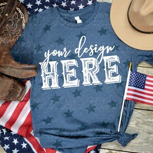 May include: A blue t-shirt with white stars and the text "your design HERE" in a vintage font. The shirt is laid out on a wooden surface with a cowboy hat, cowboy boots, and an American flag.