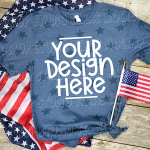 May include: A blue t-shirt with white stars printed on it. The shirt has the text "Your Design Here" printed on the front. The shirt is laid out on a wooden surface with an American flag draped over it.