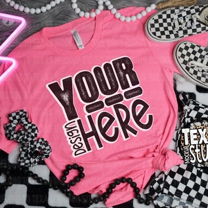 May include: Pink t-shirt with the text "Design Your Here" in black and white lettering. The shirt is laying on a black and white checkered fabric with black beads, a pink neon lightning bolt, and black and white checkered scrunchies.