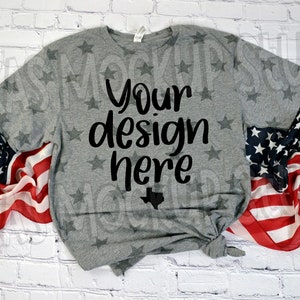 May include: Gray t-shirt with a white star pattern and the text "Your design here" in black. The t-shirt is folded and displayed on a white brick background with an American flag draped on either side.