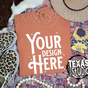 May include: A burnt orange t-shirt with the text "YOUR DESIGN HERE" in white. The t-shirt is lying on a brick background with a pair of leopard print slip-on shoes, a brown hat, and a sunflower print fabric square.