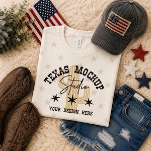 May include: A cream-colored t-shirt featuring a tan cactus graphic, black stars, and the text "TEXAS MOCKUP STUDIO YOUR DESIGN HERE". The shirt is accompanied by a distressed charcoal baseball cap with an American flag patch, brown leather cowboy boots, and blue denim shorts.
